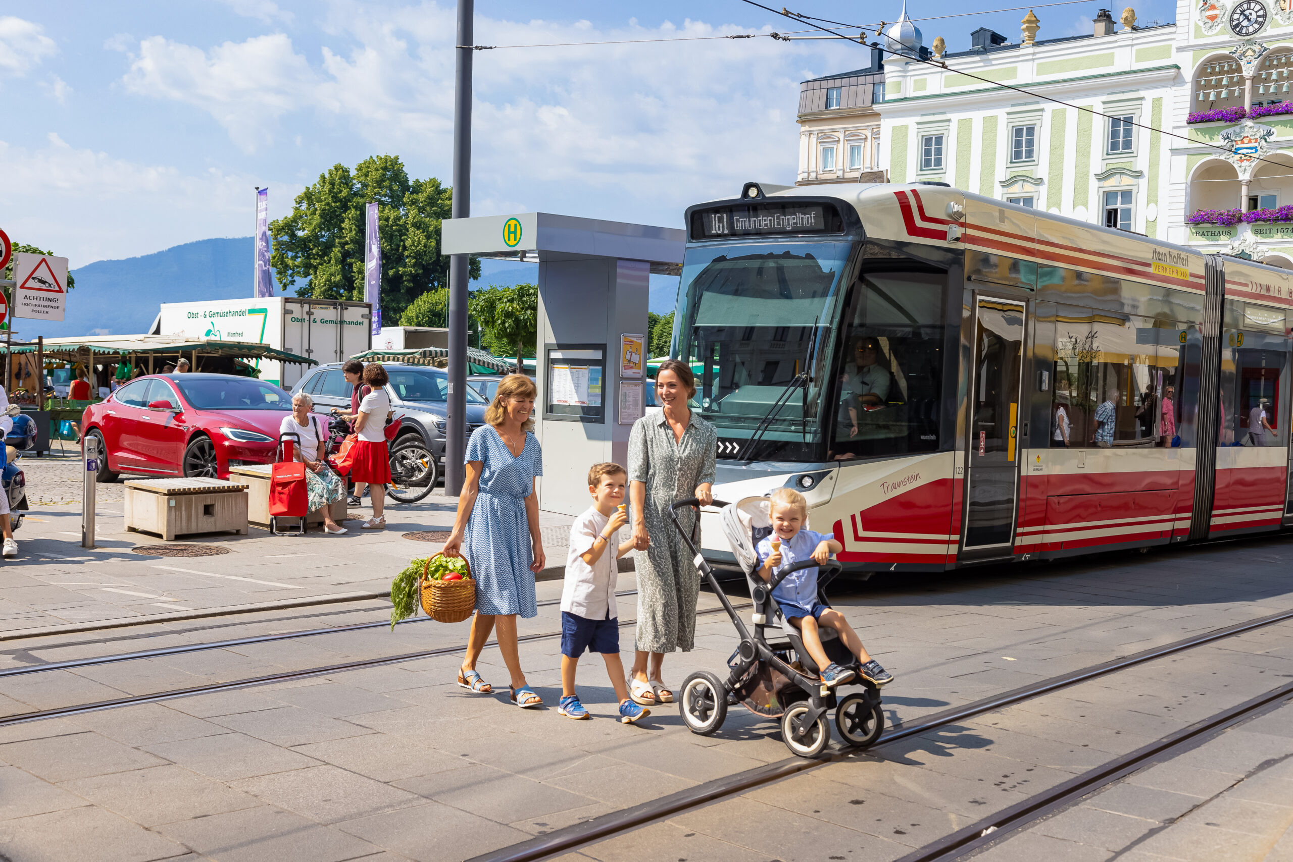 Eine Mutter geht mit iheren Kindern über die Straße vom einkaufen, die Traunseetram steht im Hintergrund